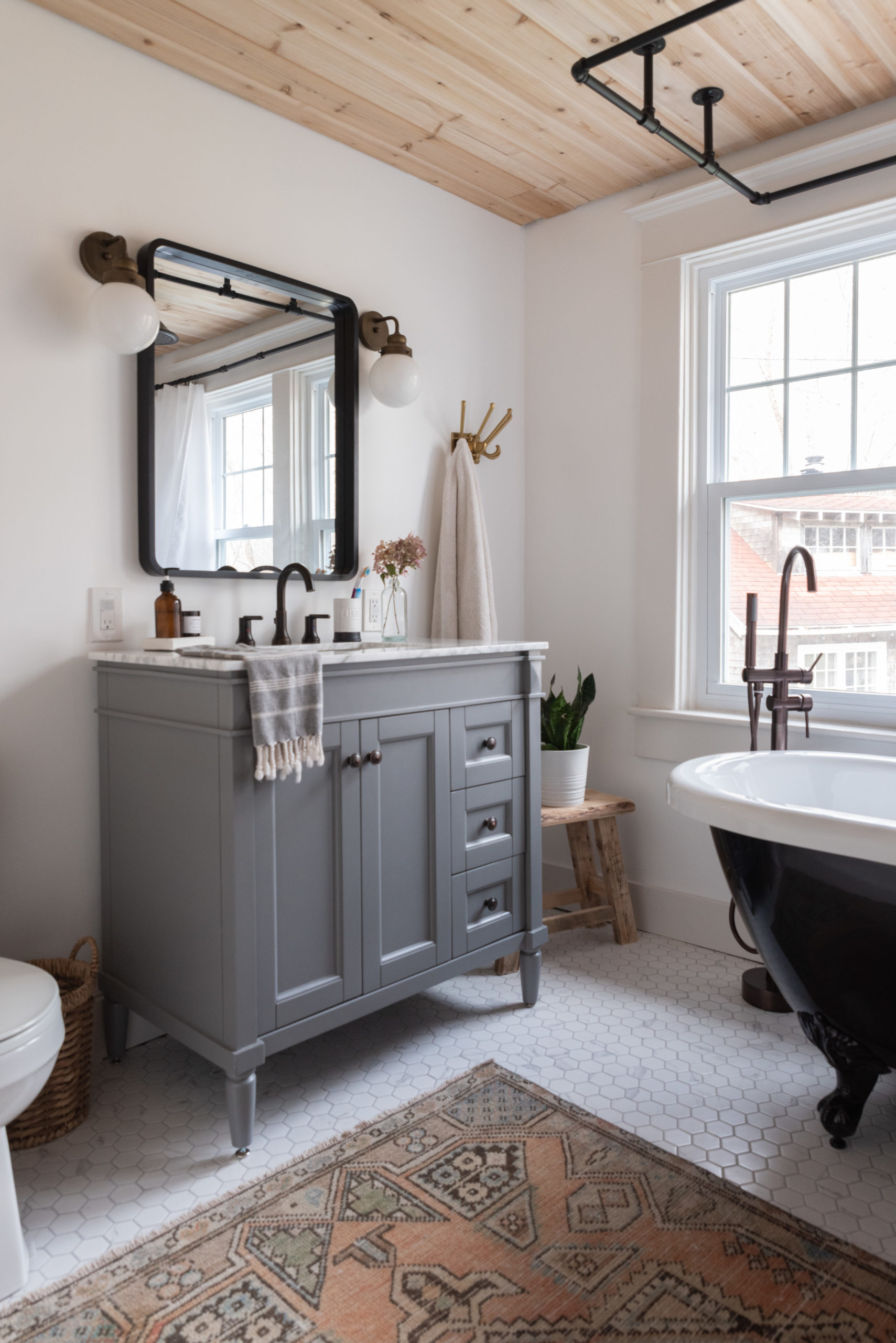 The Wild Decoelis | Our Bathroom Reno | white bathroom with gray vanity, marble hexagon floor tiles and marble countertop, black clawfoot tub, cedar plank ceiling, and an antique refinished black china cabinet used for linens