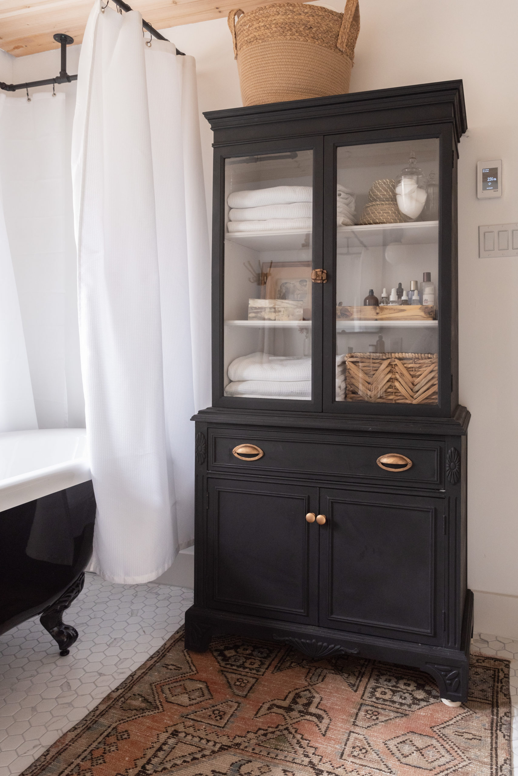 The Wild Decoelis | Our Bathroom Reno | white bathroom with gray vanity, marble hexagon floor tiles and marble countertop, black clawfoot tub, cedar plank ceiling, and an antique refinished black china cabinet used for linens