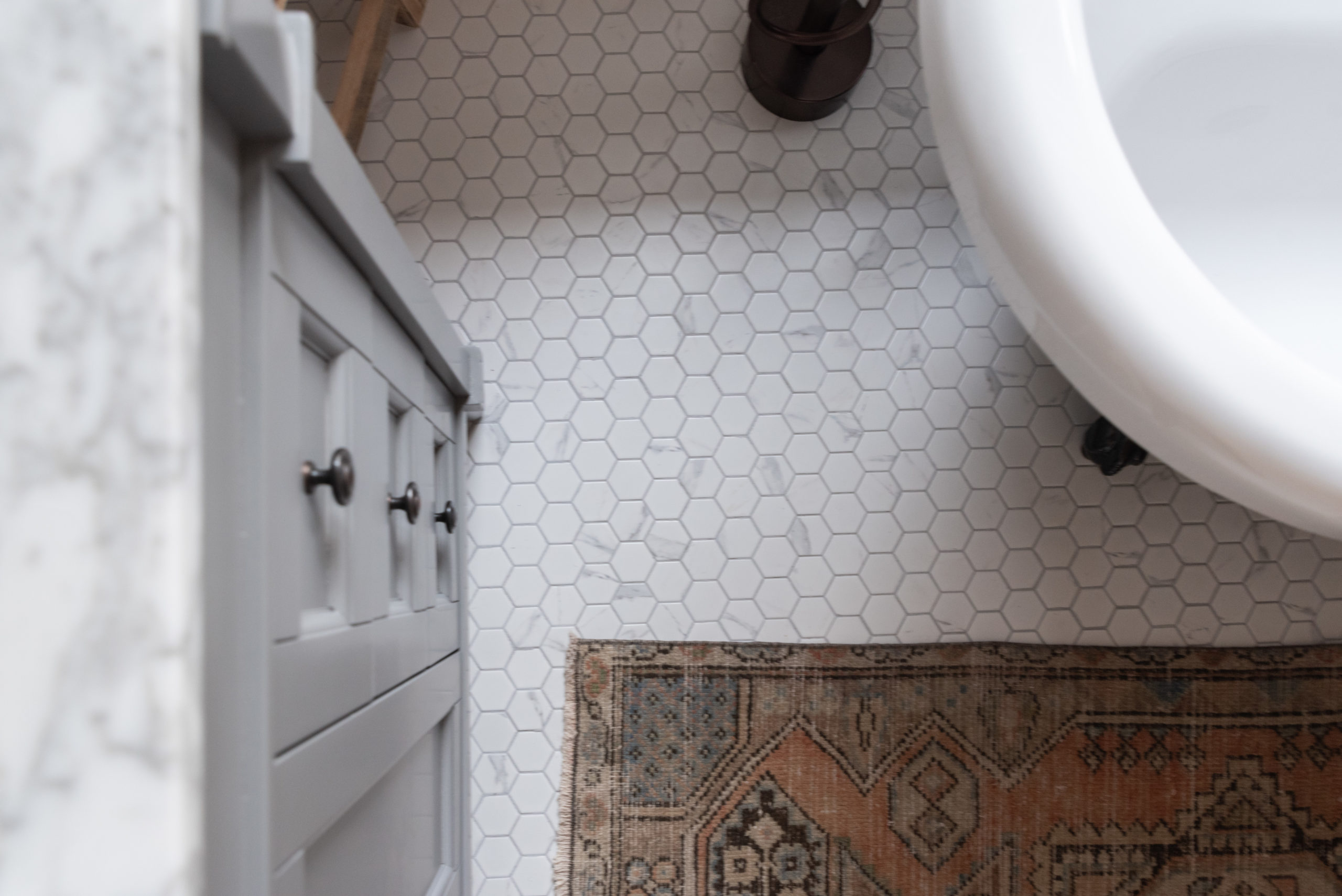 The Wild Decoelis | Our Bathroom Reno | white bathroom with gray vanity, marble hexagon floor tiles and marble countertop, black clawfoot tub, cedar plank ceiling, and an antique refinished black china cabinet used for linens vintage rug