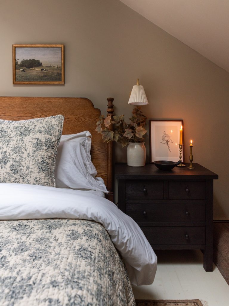 A cozy cottage-style bedroom featuring Pottery Barn wood furniture and Rejuvenation bedding in a soft floral toile pattern. The warm oak bed frame and dresser are paired with black nightstands, brass accents, and neutral walls, creating a timeless, inviting space styled with layered textiles, vintage-inspired artwork, and natural light.