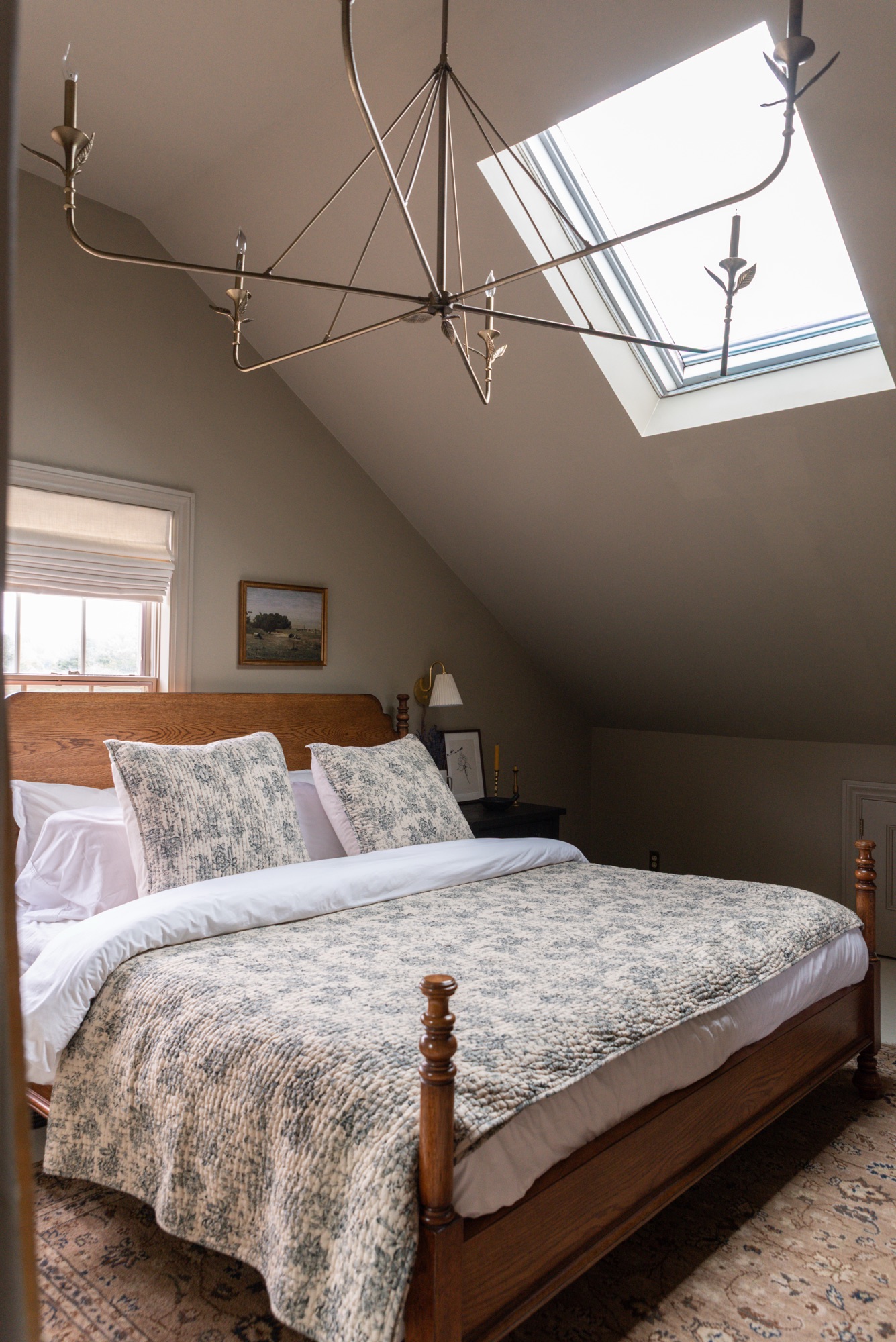 A cozy cottage-style bedroom featuring Pottery Barn wood furniture and Rejuvenation bedding in a soft floral toile pattern. The warm oak bed frame and dresser are paired with black nightstands, brass accents, and neutral walls, creating a timeless, inviting space styled with layered textiles, vintage-inspired artwork, and natural light.