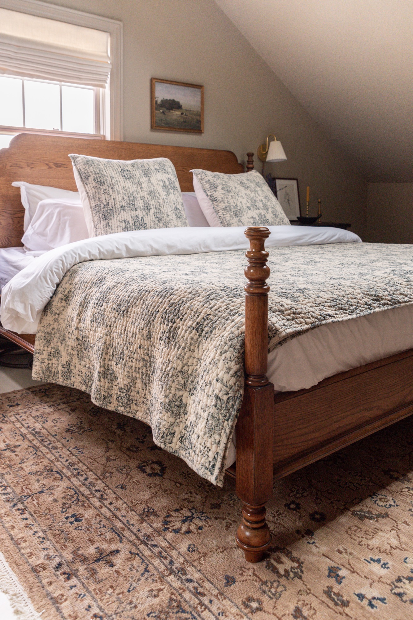 A cozy cottage-style bedroom featuring Pottery Barn wood furniture and Rejuvenation bedding in a soft floral toile pattern. The warm oak bed frame and dresser are paired with black nightstands, brass accents, and neutral walls, creating a timeless, inviting space styled with layered textiles, vintage-inspired artwork, and natural light.