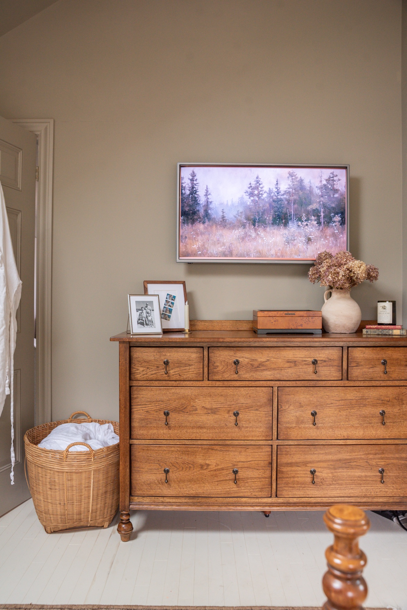 A cozy cottage-style bedroom featuring Pottery Barn wood furniture and Rejuvenation bedding in a soft floral toile pattern. The warm oak bed frame and dresser are paired with black nightstands, brass accents, and neutral walls, creating a timeless, inviting space styled with layered textiles, vintage-inspired artwork, and natural light.