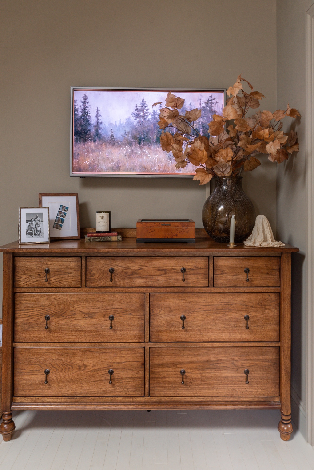 A cozy cottage-style bedroom featuring Pottery Barn wood furniture and Rejuvenation bedding in a soft floral toile pattern. The warm oak bed frame and dresser are paired with black nightstands, brass accents, and neutral walls, creating a timeless, inviting space styled with layered textiles, vintage-inspired artwork, and natural light.