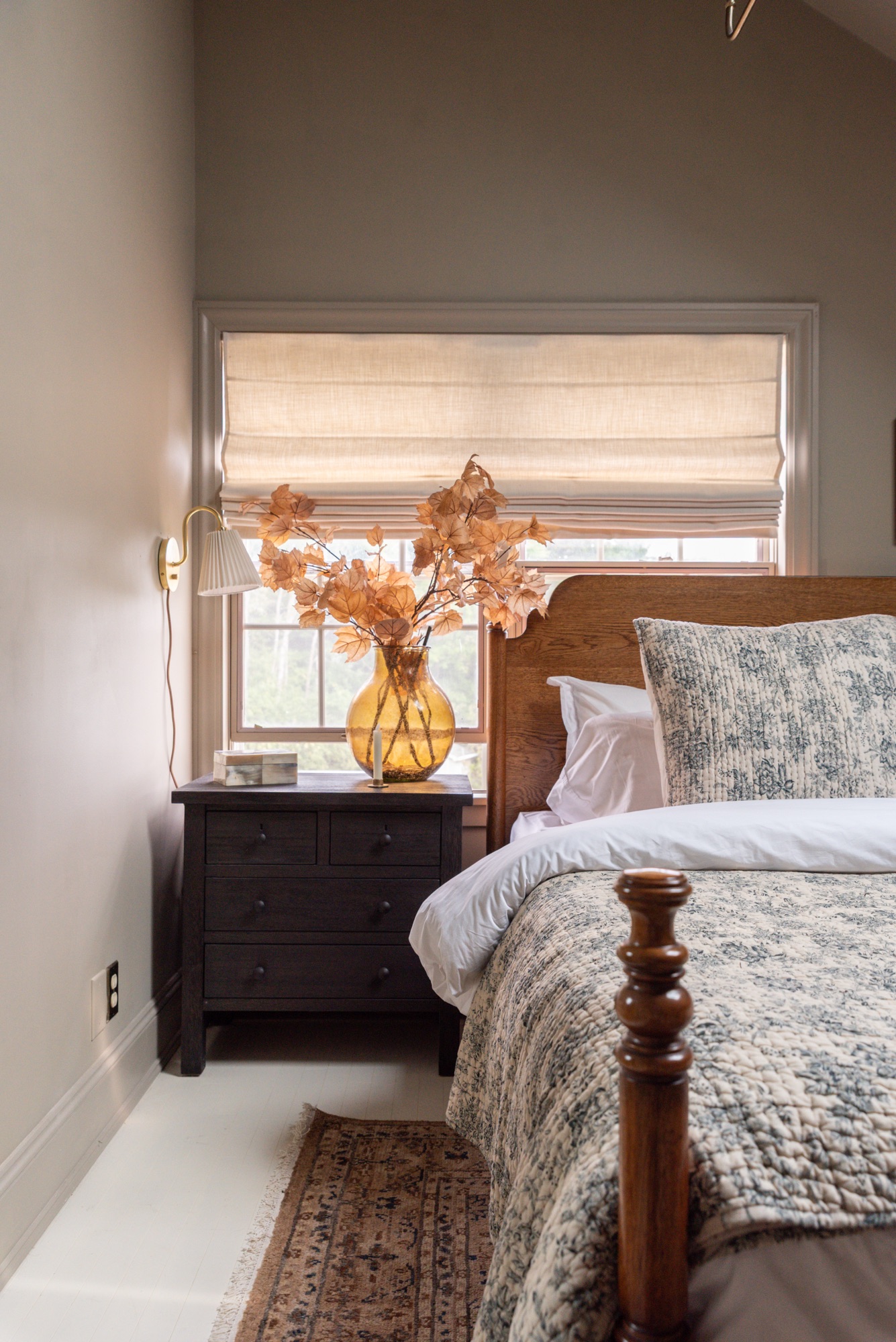 A cozy cottage-style bedroom featuring Pottery Barn wood furniture and Rejuvenation bedding in a soft floral toile pattern. The warm oak bed frame and dresser are paired with black nightstands, brass accents, and neutral walls, creating a timeless, inviting space styled with layered textiles, vintage-inspired artwork, and natural light.