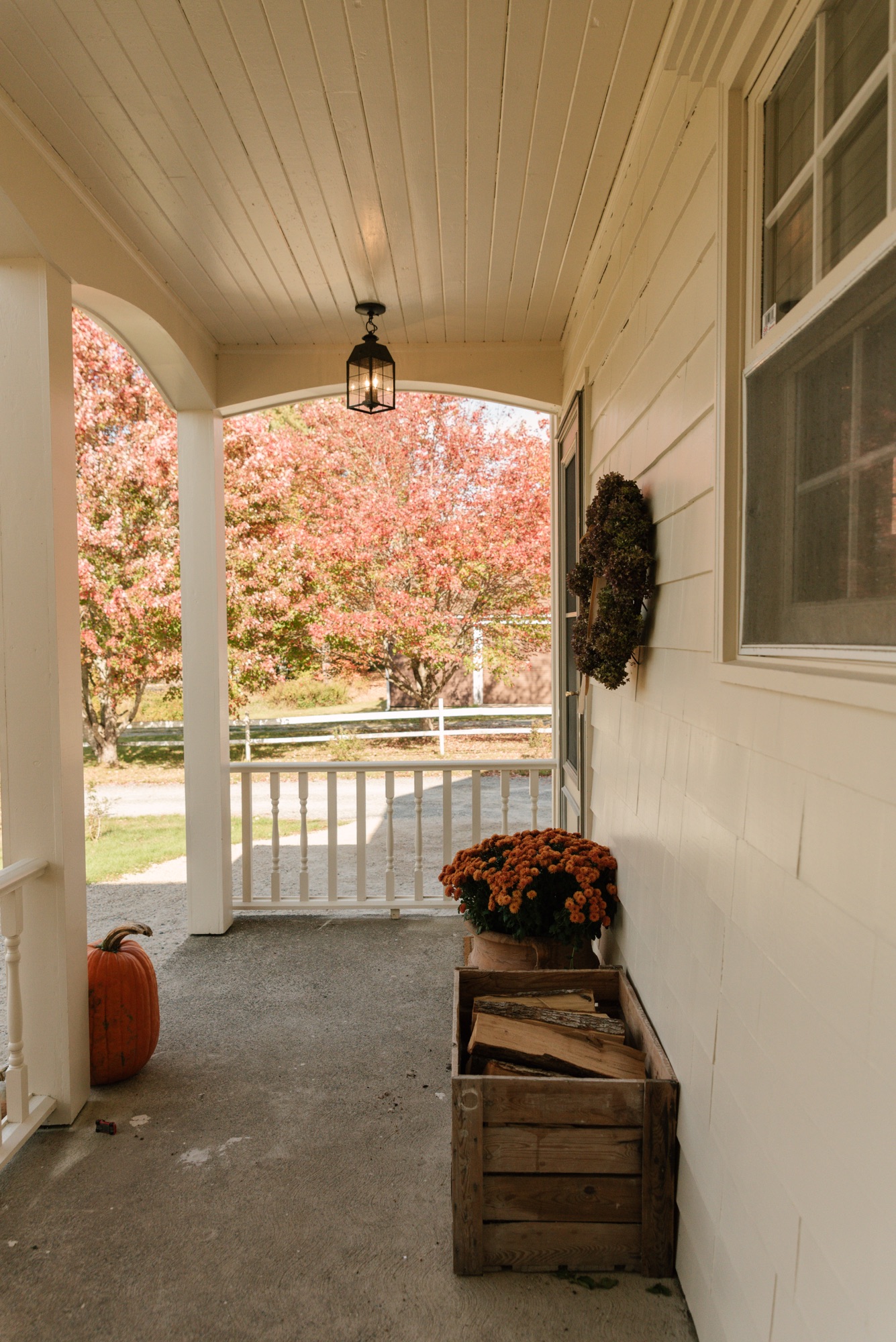 Side view of farmhouse porch with autumn trees, Hinkley lantern, and fall decorations with pumpkins and mums.